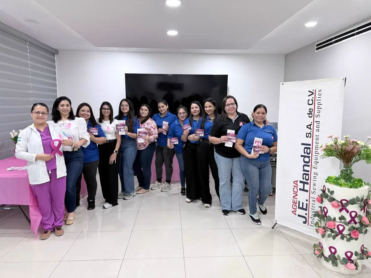 Colaboradoras de Agencia J.E. Handal en el showroom de la empresa, con material informativo sobre el cáncer de mama y un lazo rosa, junto a la doctora Vanessa Argueta, que impartió la charla